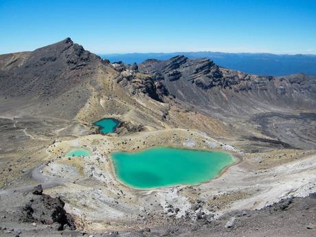 A view of emerald-green lakes nestled among barren volcanic terrain, with rugged mountains in the background.