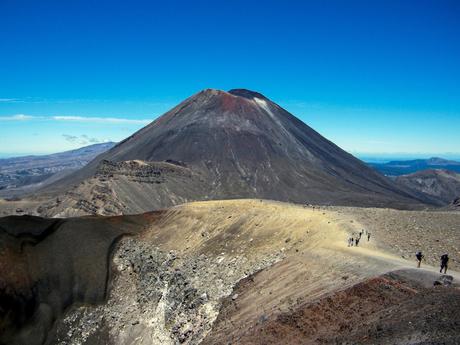 A volcanic mountain peak under a bright blue sky, with a trail winding up towards the summit and a group of hikers in the distance.