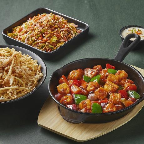 A sizzling pan of chilli paneer, a vibrant bowl of Chinese bhel bursting with mixed vegetables, and a platter of crispy toppings to add on your bhel.