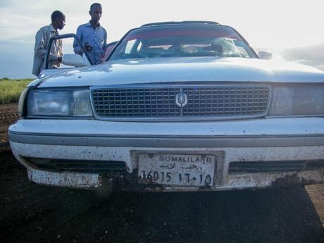 A close-up shot of a white car with a muddy front and a Somaliland license plate, two men standing beside it.