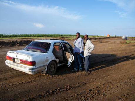 Two men stand beside an old white sedan on a rough dirt road in Somaliland, with fields stretching out in the background.