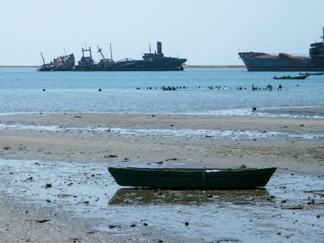 A sandy beach with a few old, rusting shipwrecks in the water and a small green boat on the shore in Somaliland.