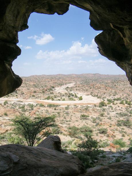 A scenic view from within a cave, looking out over a rugged landscape of rocky terrain and sparse vegetation under a partly cloudy sky.