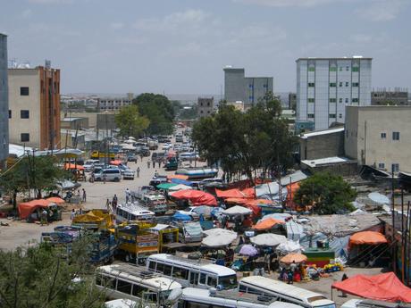 A bustling street market in a city in Somaliland, filled with vendors, vehicles, and people under bright awnings.