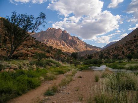 A scenic mountain landscape with a dirt path and a small stream running through a grassy valley under a partly cloudy sky.