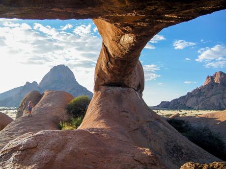 A rock arch frames a view of a rocky desert landscape with a person standing on a boulder and a large mountain in the distance.