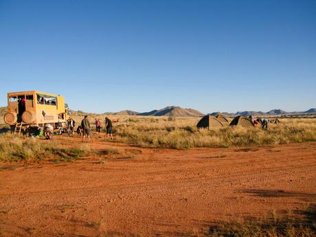 A group of travelers setting up camp next to a large, rugged off-road vehicle in a flat, grassy plain under a clear blue sky.