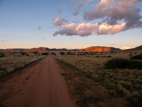 A dirt road stretches towards distant hills illuminated by the warm light of the setting sun, with a partly cloudy sky above.