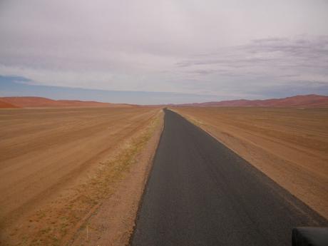 A long, straight road cutting through a vast, arid desert landscape, with reddish dunes visible in the distance under a cloudy sky.