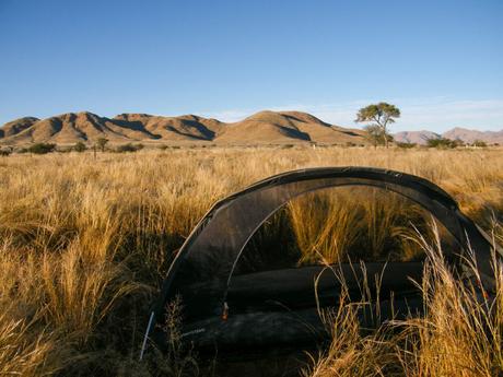 A small see-through freestanding mosquito net tent set up in a grassy field with rugged hills in the background under a clear blue sky.