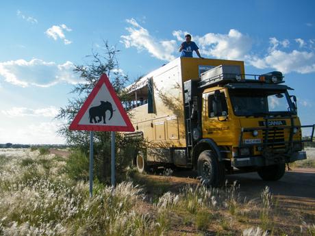 A yellow overland truck parked beside a sign warning of elephants, with a man standing on top of the truck waving.