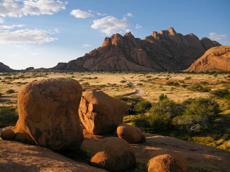 Large boulders in the foreground with a dirt path leading toward a dramatic, rocky mountain range under a clear blue sky.