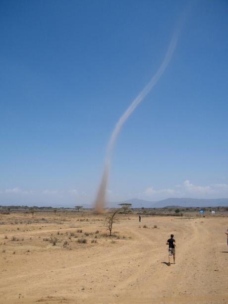 A dust devil spiraling into the clear blue sky over a dry, barren landscape, with small figures in the distance.