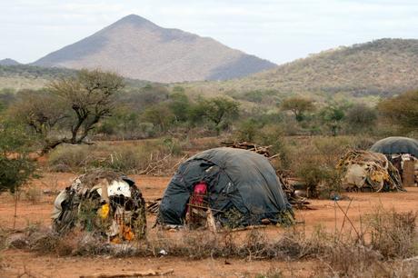 Several makeshift shelters made from tarps and natural materials in a rural landscape, with a backdrop of hills and dry, bushy vegetation.