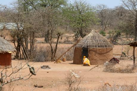 A traditional circular mud hut with a thatched roof in a rural village, with a woman in a yellow dress crouching near the entrance, surrounded by dry, sparse vegetation.