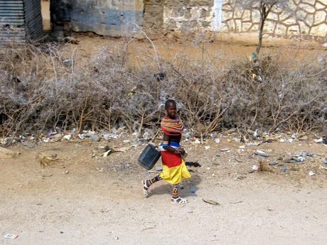 A young person wearing a colorful traditional dress walks along a dusty roadside, carrying a black plastic container, with dry bushes and stone walls in the background.