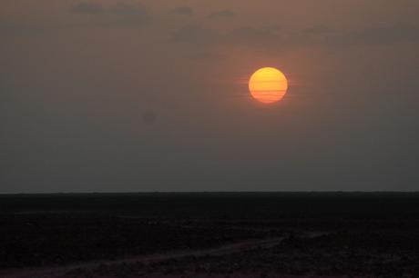 A large orange sun setting over a dark, flat landscape, with the horizon and sky blending into soft hues of pink and gray.