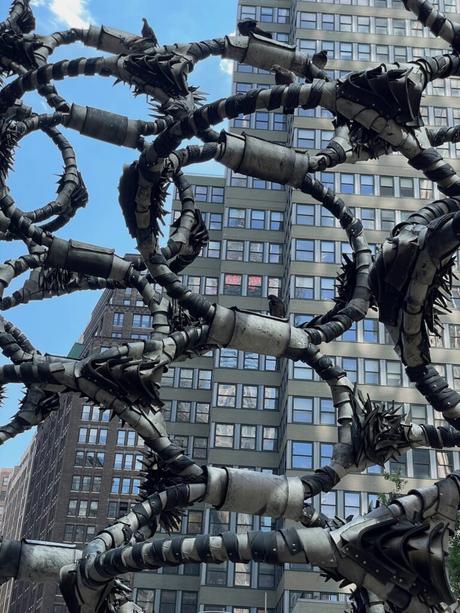 A unique metal sculpture of twisted, spiked tendrils in a public plaza with tall buildings in the background.