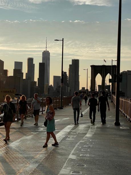 People walking on the Brooklyn Bridge at sunset, with One World Trade Center and other skyscrapers of downtown Manhattan in the distance.