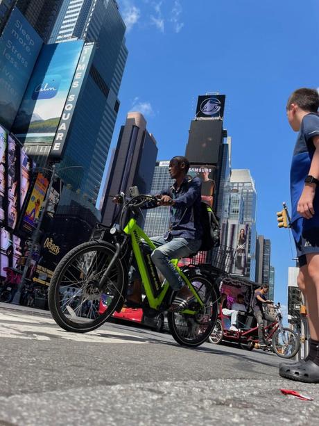 A cyclist riding a bright green electric bike through Times Square, with large billboards and tall buildings in the background.