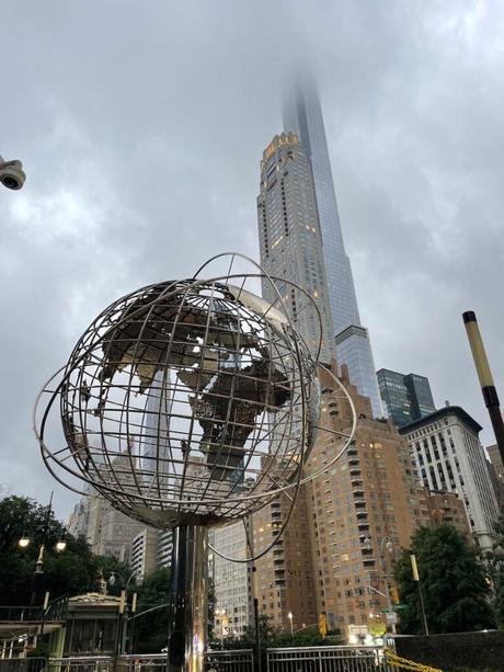 A metal globe sculpture in front of skyscrapers in New York City, with the tallest building partially obscured by fog or clouds.