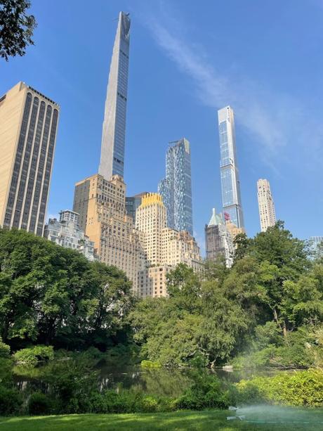 A view of Central Park with a small pond in the foreground and New York City's towering skyscrapers, including the ultra-tall 432 Park Avenue, rising in the background. The lush greenery contrasts with the modern skyline under a clear blue sky.