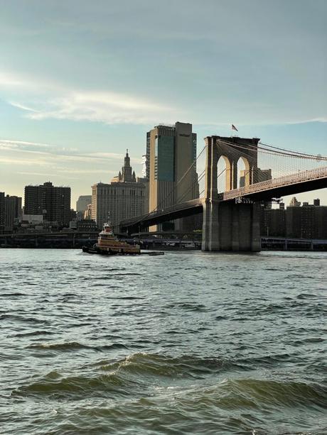 The Brooklyn Bridge at sunset, with a tugboat on the water and downtown Manhattan's skyline in the background.