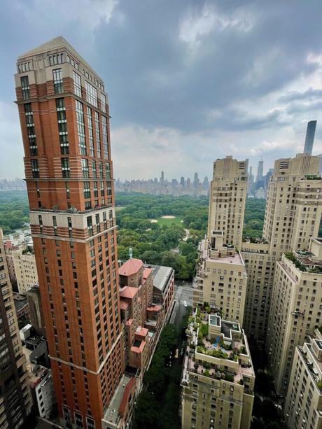 A view from a high-rise in New York City showing a tall, red-brick residential building and other skyscrapers surrounding Central Park. The park's lush green trees spread out beneath a cloudy sky, with the city's skyline visible in the distance.