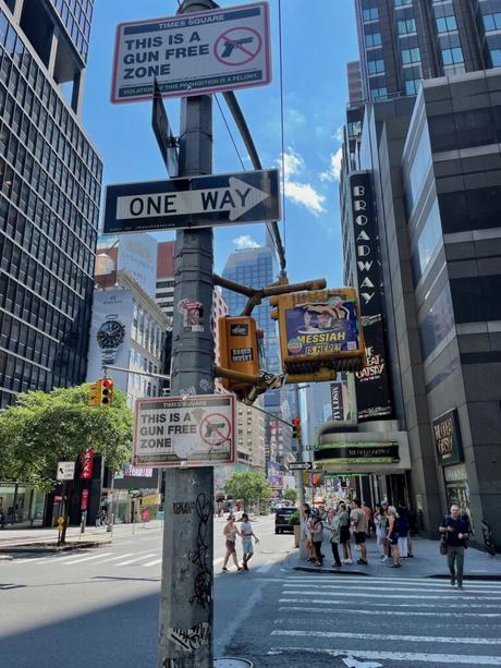 A street sign in Times Square declaring the area a 'Gun Free Zone' with Broadway signs and a busy intersection in the background.