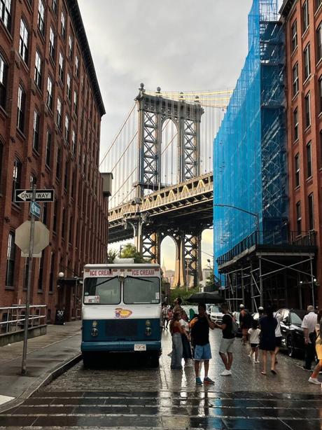 A view of the Manhattan Bridge framed by red brick buildings in the Dumbo neighborhood of Brooklyn, with an ice cream truck parked on the street.