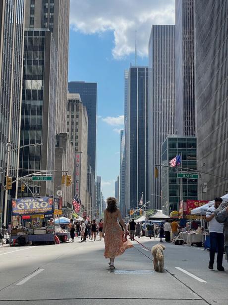 A woman in a floral dress walking her dog along a closed-off New York City street, surrounded by skyscrapers and food stalls.