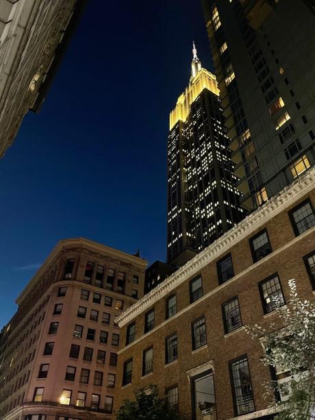 A nighttime view of the Empire State Building illuminated in yellow, framed by surrounding buildings under a dark blue sky.