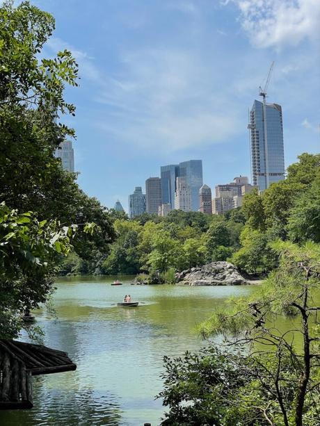 A scenic view of a small lake in Central Park, New York City, with rowboats gliding across the water. Lush green trees frame the lake, while the city's modern skyscrapers rise in the distance under a sunny sky.