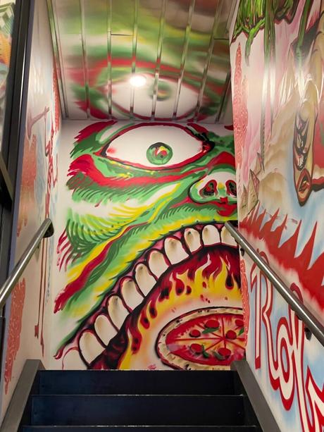 A brightly colored mural inside a staircase, depicting a green, red, and yellow stylized face with an open mouth, flames, and a pizza.