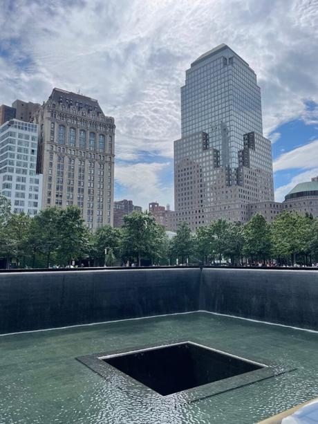 The 9/11 Memorial South Pool at Ground Zero, New York City, featuring a large square waterfall surrounded by trees and skyscrapers under a partly cloudy sky.