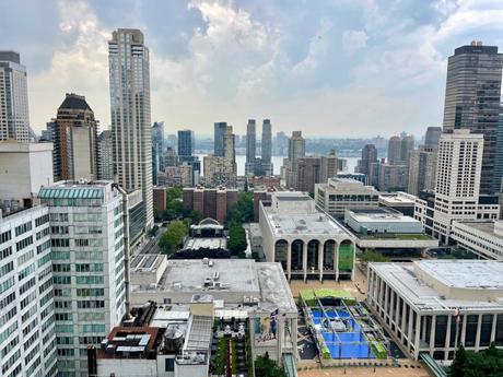 Aerial view of Lincoln Center in New York City, featuring modern buildings surrounding a central plaza with a blue event setup. Skyscrapers and residential towers stretch toward the Hudson River in the distance under a cloudy sky.