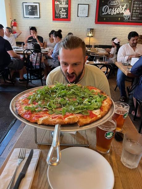 Alex Tiffany humorously preparing to eat a large arugula-topped pizza at a restaurant in New York City, with other diners seated at tables in the background.