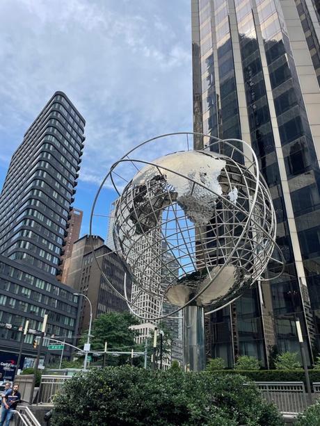 A large metal globe sculpture standing in front of modern skyscrapers in Columbus Circle, New York City, under a partly cloudy sky.