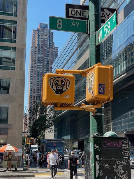 A street sign at the corner of 8th Avenue and 58th Street in New York City, with a pedestrian crossing signal featuring a quirky dog sticker.