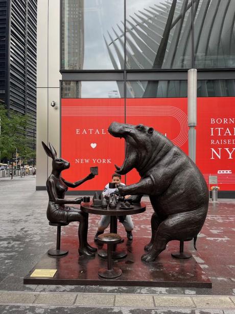 A whimsical bronze sculpture of a rabbit and a hippo sitting at a table enjoying coffee outside the Eataly market in New York City, with a young boy posing behind the sculpture.