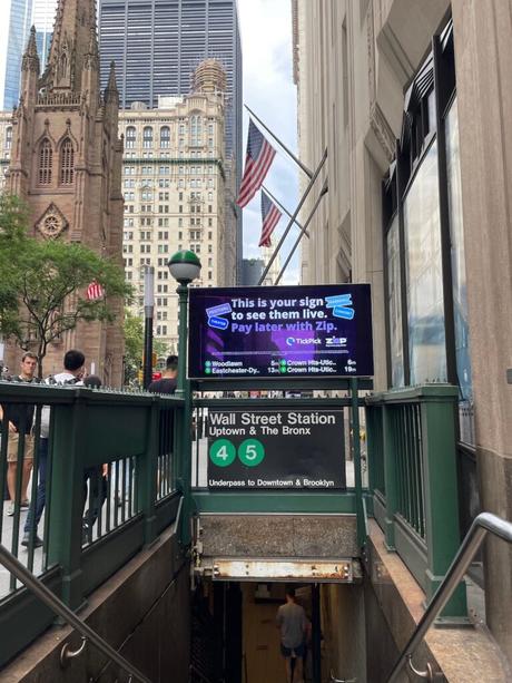 The entrance to Wall Street subway station in New York City, with the iconic green sign and American flags displayed above. In the background, Trinity Church is visible among the tall buildings.