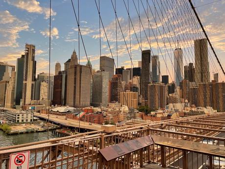 The New York City skyline at sunset, viewed from the Brooklyn Bridge, with skyscrapers framed by suspension cables and a colorful sky.