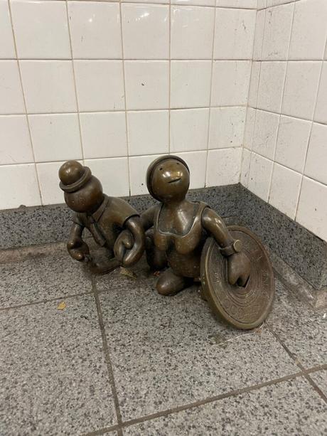 A small bronze sculpture of two cartoonish figures, one holding a giant coin, at a New York City subway station.