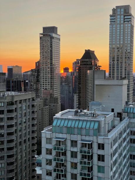 Sunset over New York City’s skyline, viewed from a high-rise, with the sun setting between tall buildings and reflecting off their windows.