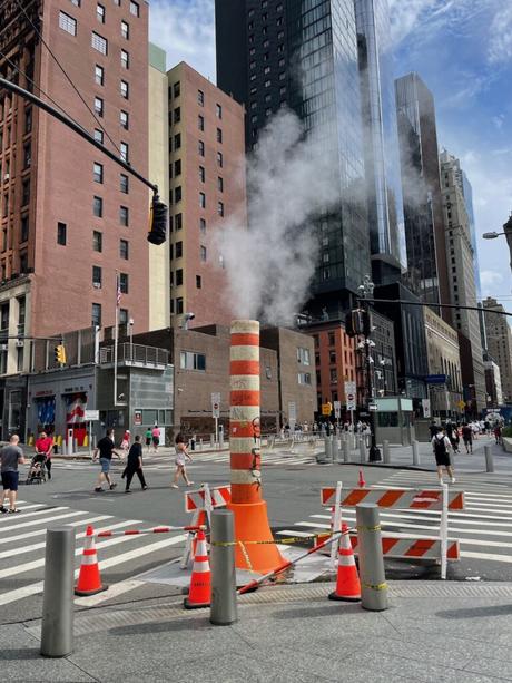 A street scene in New York City showing a steam pipe emitting vapor through a striped orange and white column, surrounded by traffic cones and construction barriers. Pedestrians walk along the crosswalk, with tall buildings and skyscrapers in the background under a partly cloudy sky.