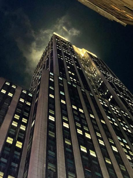 A close-up, upward angle of the Empire State Building at night, with clouds surrounding the illuminated tower.