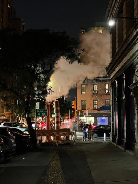 Steam rising from a construction pipe at night on a New York City street, with cars and buildings lit by streetlights.