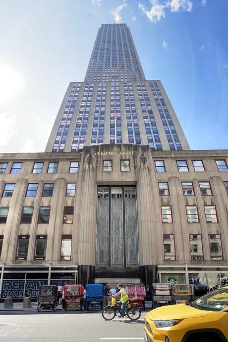 A low-angle view of the Empire State Building with its Art Deco facade and cyclists riding past yellow taxis on the street below.