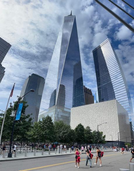 The One World Trade Center skyscraper reflecting the sky, towering over nearby buildings and trees, with pedestrians walking along the street below.