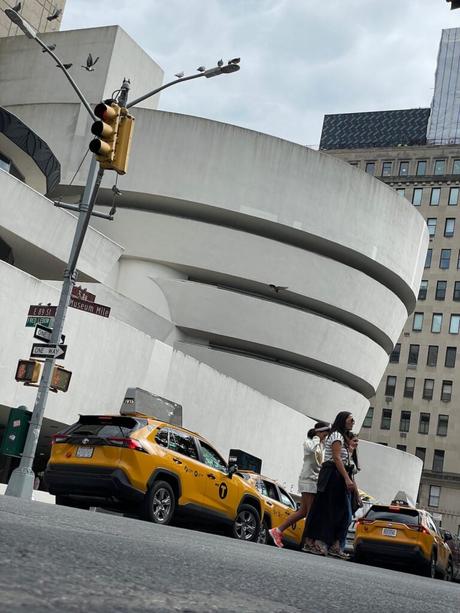 Yellow taxis in front of the Guggenheim Museum in New York, with people walking by on the sidewalk and pigeons perched on a streetlight.